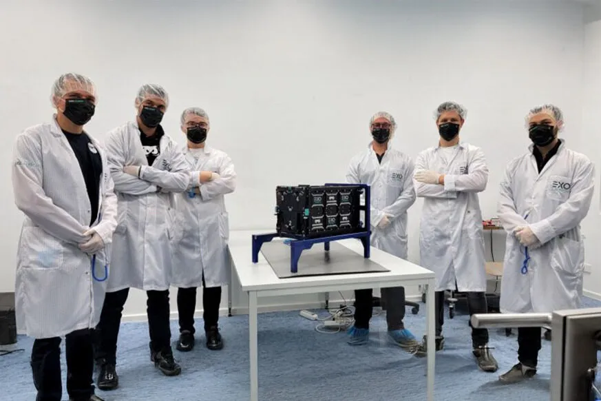 A team of six engineers and scientists wearing white lab coats, black face masks, hairnets, and gloves stand in a cleanroom around a small satellite mounted on a blue test stand. The satellite, covered in black panels with multiple connectors and components, is placed on a white table in a controlled environment. Some team members have their arms crossed, while others hold tools, signifying the final preparations before launch. The EXO logo is visible on their lab coats, indicating a collaboration with Exolaunch. The setting reflects a high-tech aerospace lab, emphasizing precision and innovation in satellite deployment.