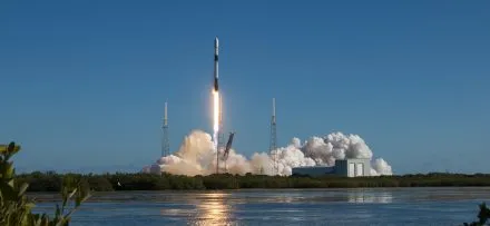 A Falcon 9 rocket lifts off from its launch pad under a clear blue sky, leaving behind a fiery trail and a billowing cloud of smoke. The launch is captured from a distance, with a reflective body of water in the foreground and lush greenery framing the scene.