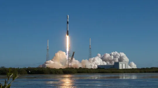 A Falcon 9 rocket lifts off from its launch pad under a clear blue sky, leaving behind a fiery trail and a billowing cloud of smoke. The launch is captured from a distance, with a reflective body of water in the foreground and lush greenery framing the scene.