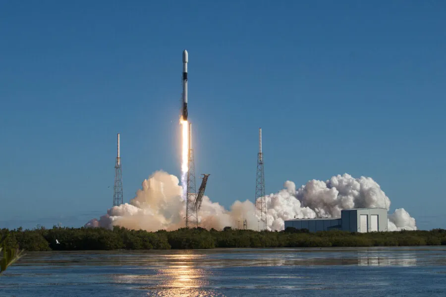 A Falcon 9 rocket lifts off from its launch pad under a clear blue sky, leaving behind a fiery trail and a billowing cloud of smoke. The launch is captured from a distance, with a reflective body of water in the foreground and lush greenery framing the scene.