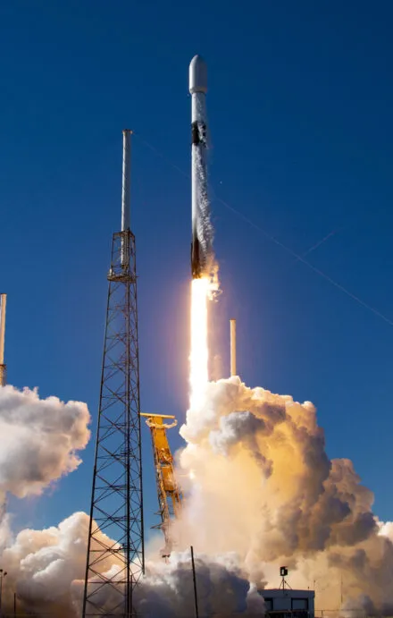 A Falcon 9 rocket ignites and lifts off into the clear blue sky, leaving behind a trail of fire and thick white smoke. The powerful launch marks another milestone in space exploration and satellite deployment, with the booster ascending rapidly above the launch pad, supported by towering metal structures.