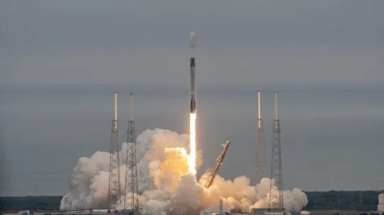A Falcon 9 rocket lifts off from its launch pad, producing a bright trail of fire and thick plumes of smoke as it ascends into the sky. The launch infrastructure, including four tall support towers, surrounds the rocket as it departs over the ocean in the background. The scene captures the power and precision of the SpaceX launch, likely carrying a payload such as satellites for Unseenlabs or another mission dedicated to space-based intelligence and communications. The moment reflects technological achievement and the advancement of satellite capabilities in maritime surveillance and security.