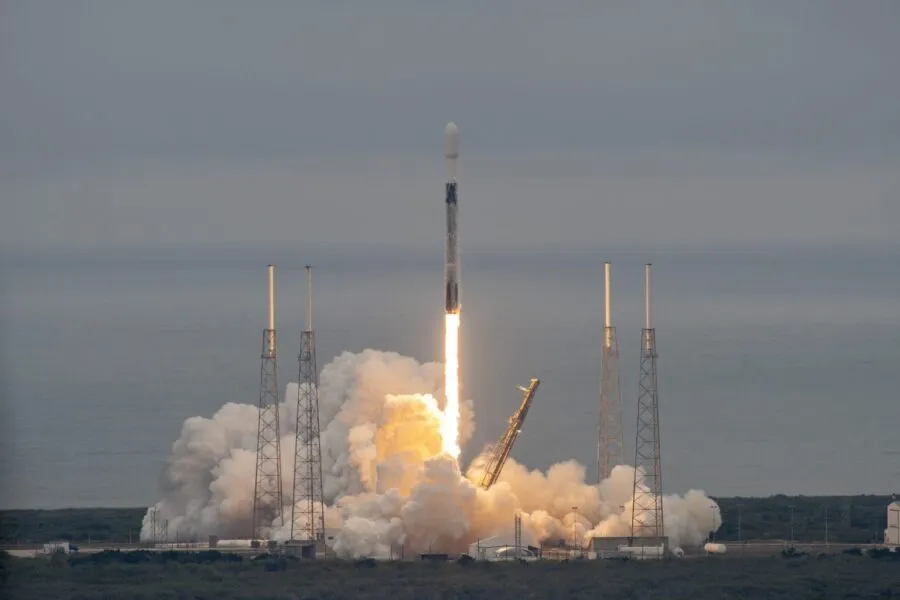 A Falcon 9 rocket lifts off from its launch pad, producing a bright trail of fire and thick plumes of smoke as it ascends into the sky. The launch infrastructure, including four tall support towers, surrounds the rocket as it departs over the ocean in the background. The scene captures the power and precision of the SpaceX launch, likely carrying a payload such as satellites for Unseenlabs or another mission dedicated to space-based intelligence and communications. The moment reflects technological achievement and the advancement of satellite capabilities in maritime surveillance and security.