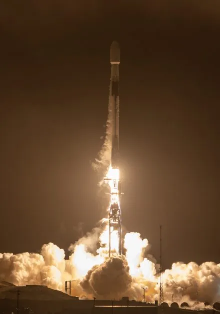A Falcon 9 rocket lifts off into the night sky, illuminating the launch pad with a fiery glow. The rocket, surrounded by thick clouds of smoke and vapor, ascends rapidly, marking another successful mission.