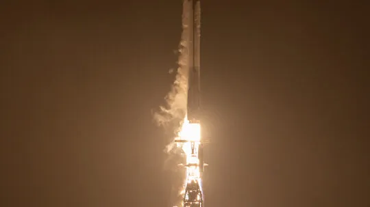 A Falcon 9 rocket lifts off into the night sky, illuminating the launch pad with a fiery glow. The rocket, surrounded by thick clouds of smoke and vapor, ascends rapidly, marking another successful mission.
