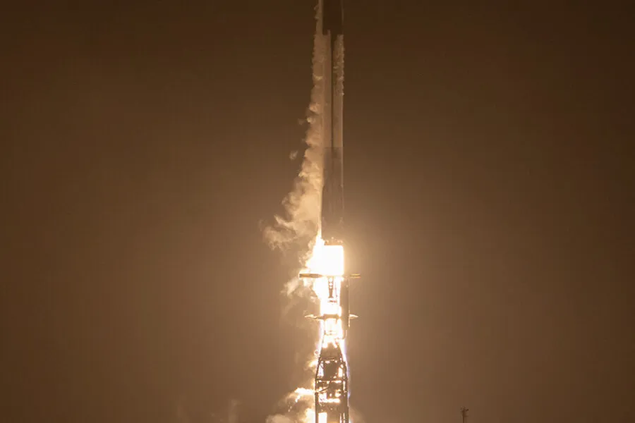 A Falcon 9 rocket lifts off into the night sky, illuminating the launch pad with a fiery glow. The rocket, surrounded by thick clouds of smoke and vapor, ascends rapidly, marking another successful mission.