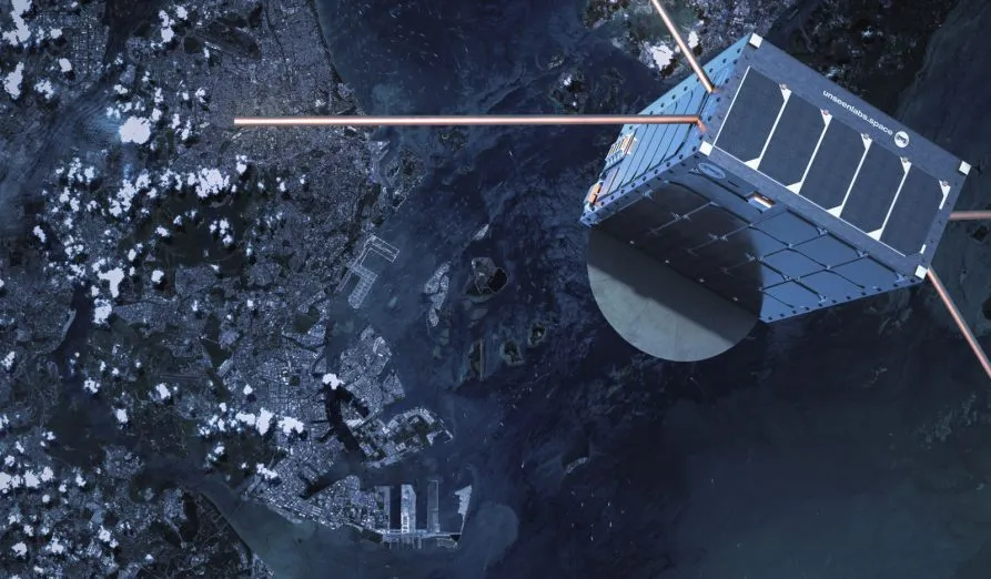 A high-resolution image of an Unseenlabs satellite orbiting Earth, showcasing its cubic design, solar panels, and extended antennas for radio frequency (RF) detection. The satellite is positioned above a coastal city with harbors and waterways visible below, emphasizing its role in maritime surveillance. The satellite's shadow is cast on its metallic surface, while the Earth’s surface, scattered with clouds, provides a striking contrast. This image highlights Unseenlabs’ space-based intelligence capabilities in tracking and monitoring vessels across global waters.