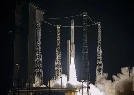 A Vega rocket, operated by the European Space Agency (ESA), is captured mid-launch at night, ascending into the dark sky with a bright plume of fire and smoke. The launchpad is surrounded by tall metal structures and cables, with the ESA and Vega logos visible on the rocket and nearby infrastructure. The scene is illuminated by the intense glow of the rocket's propulsion, highlighting the dramatic moment of liftoff.