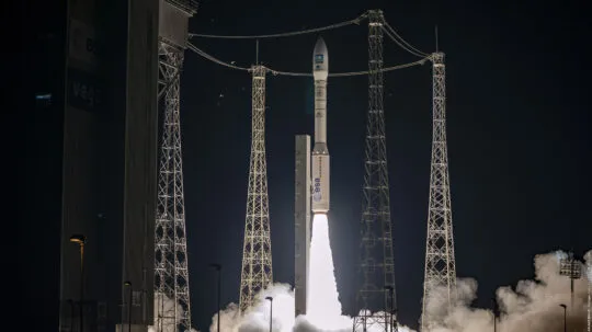 A Vega rocket, operated by the European Space Agency (ESA), is captured mid-launch at night, ascending into the dark sky with a bright plume of fire and smoke. The launchpad is surrounded by tall metal structures and cables, with the ESA and Vega logos visible on the rocket and nearby infrastructure. The scene is illuminated by the intense glow of the rocket's propulsion, highlighting the dramatic moment of liftoff.