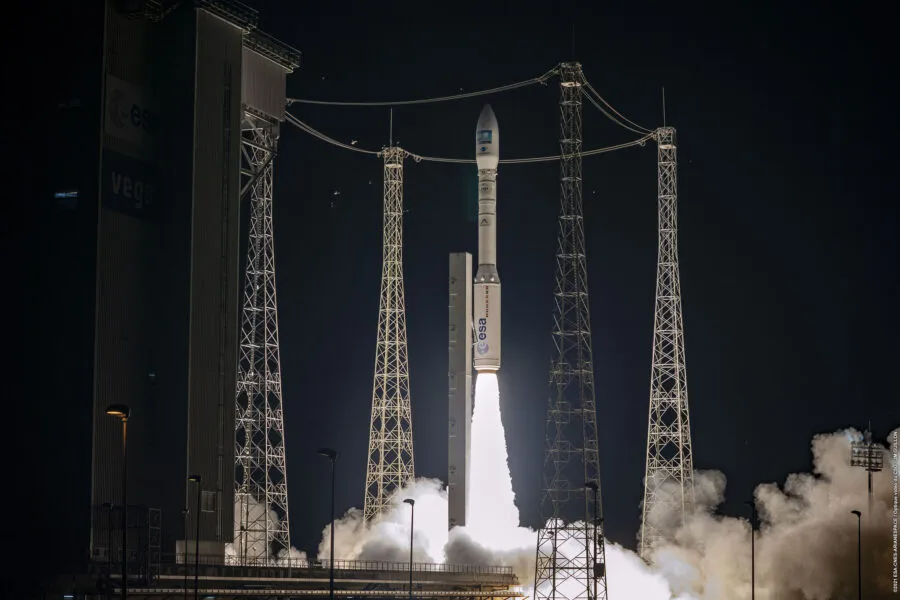 A Vega rocket, operated by the European Space Agency (ESA), is captured mid-launch at night, ascending into the dark sky with a bright plume of fire and smoke. The launchpad is surrounded by tall metal structures and cables, with the ESA and Vega logos visible on the rocket and nearby infrastructure. The scene is illuminated by the intense glow of the rocket's propulsion, highlighting the dramatic moment of liftoff.