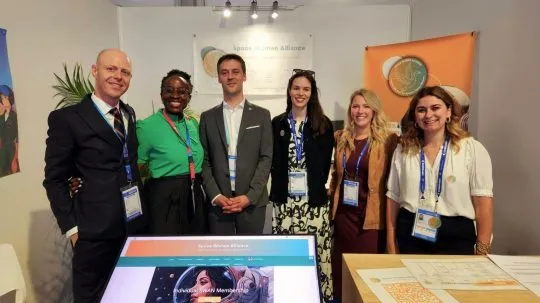 Group of six professionals, from left to right : Rolland.Z , Pamela.M, Clément.G, Mélanie.F, Cannelle.G & Cécile.R, smiling at the Space Women Alliance booth during a space industry event, standing in front of banners and promotional materials.