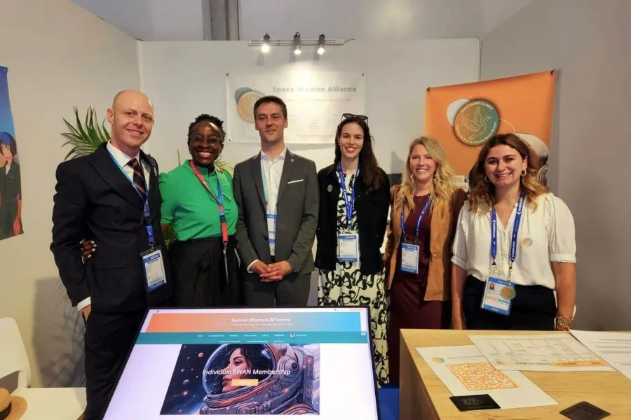 Group of six professionals, from left to right : Rolland.Z , Pamela.M, Clément.G, Mélanie.F, Cannelle.G & Cécile.R, smiling at the Space Women Alliance booth during a space industry event, standing in front of banners and promotional materials.