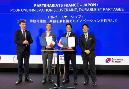 Quatre hommes en costume posent sur scène lors d’une cérémonie de signature autour d’un partenariat franco-japonais. Au centre, deux d’entre eux tiennent des dossiers ouverts devant une table avec de petits drapeaux français, européen et japonais. En arrière-plan, un grand écran bleu affiche en français et en japonais : « Partenariats France-Japon : pour une innovation souveraine, durable et partagée », avec les logos de Business France et de la République française.