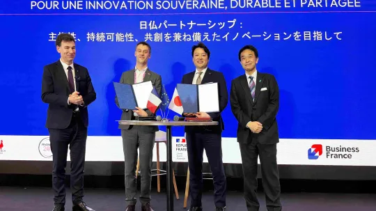 Quatre hommes en costume posent sur scène lors d’une cérémonie de signature autour d’un partenariat franco-japonais. Au centre, deux d’entre eux tiennent des dossiers ouverts devant une table avec de petits drapeaux français, européen et japonais. En arrière-plan, un grand écran bleu affiche en français et en japonais : « Partenariats France-Japon : pour une innovation souveraine, durable et partagée », avec les logos de Business France et de la République française.