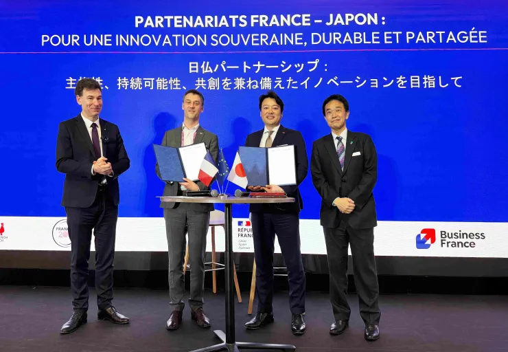 Quatre hommes en costume posent sur scène lors d’une cérémonie de signature autour d’un partenariat franco-japonais. Au centre, deux d’entre eux tiennent des dossiers ouverts devant une table avec de petits drapeaux français, européen et japonais. En arrière-plan, un grand écran bleu affiche en français et en japonais : « Partenariats France-Japon : pour une innovation souveraine, durable et partagée », avec les logos de Business France et de la République française.