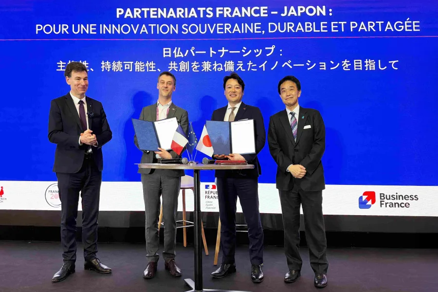 Quatre hommes en costume posent sur scène lors d’une cérémonie de signature autour d’un partenariat franco-japonais. Au centre, deux d’entre eux tiennent des dossiers ouverts devant une table avec de petits drapeaux français, européen et japonais. En arrière-plan, un grand écran bleu affiche en français et en japonais : « Partenariats France-Japon : pour une innovation souveraine, durable et partagée », avec les logos de Business France et de la République française.